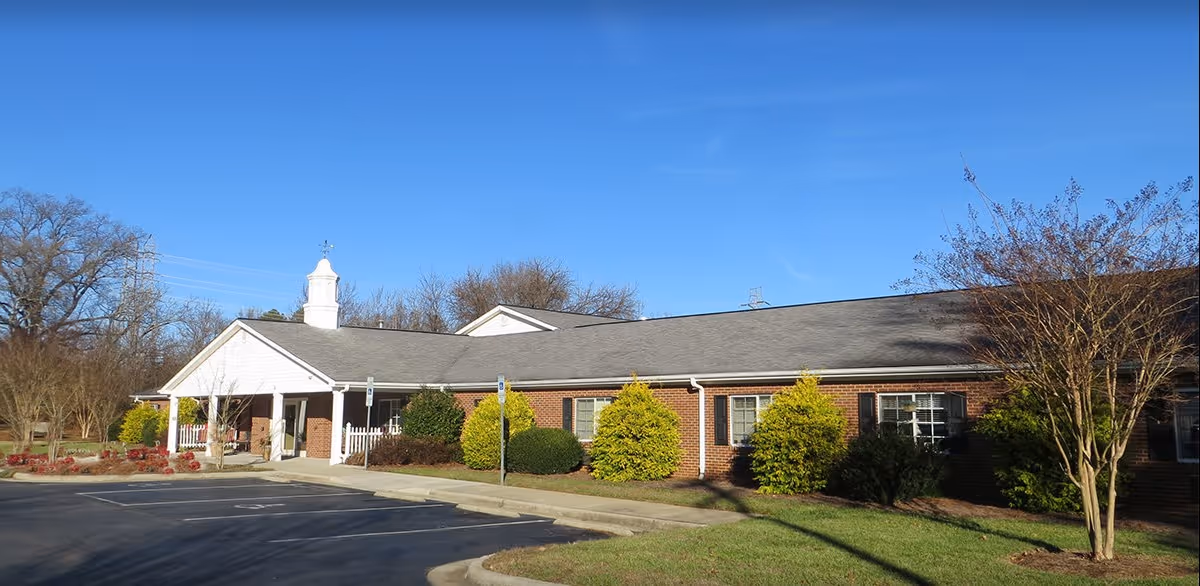 Exterior view of a single-story brick building with a gray roof and white trim, surrounded by small bushes and trees under a clear blue sky. There is a paved parking lot in front with marked parking spaces.