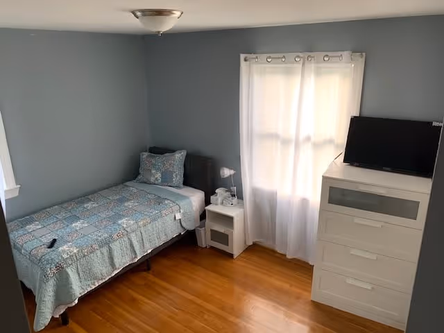 A small bedroom with light blue walls and hardwood floors. There is a single bed with a blue patterned quilt and matching pillow against one wall. Next to the bed is a white nightstand with a lamp and some items on it. A white dresser with a small flat-screen TV on top is positioned near a window with white curtains.