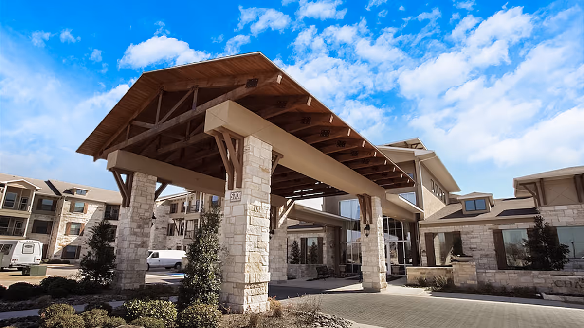 Exterior view of Chateau McKinney senior living facility showing a large covered entrance with stone pillars and wooden beams under a bright blue sky with scattered clouds.