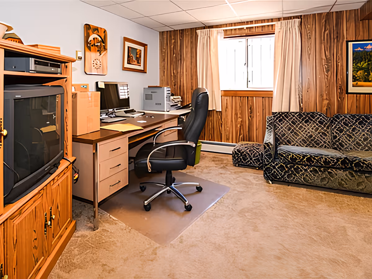 A cozy room with wood-paneled walls and beige carpet featuring a black office chair on a plastic mat in front of a desk with a computer and printer. To the left is a wooden TV cabinet with an older-style television, and to the right is a dark patterned couch under a window with beige curtains.