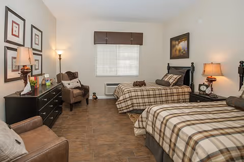 A cozy bedroom with two twin beds featuring plaid bedding. The room includes a dark wooden dresser with a lamp and decorative items, a comfortable armchair, a floor lamp, and a window with blinds and a valance. The walls are light-colored and decorated with framed artwork.