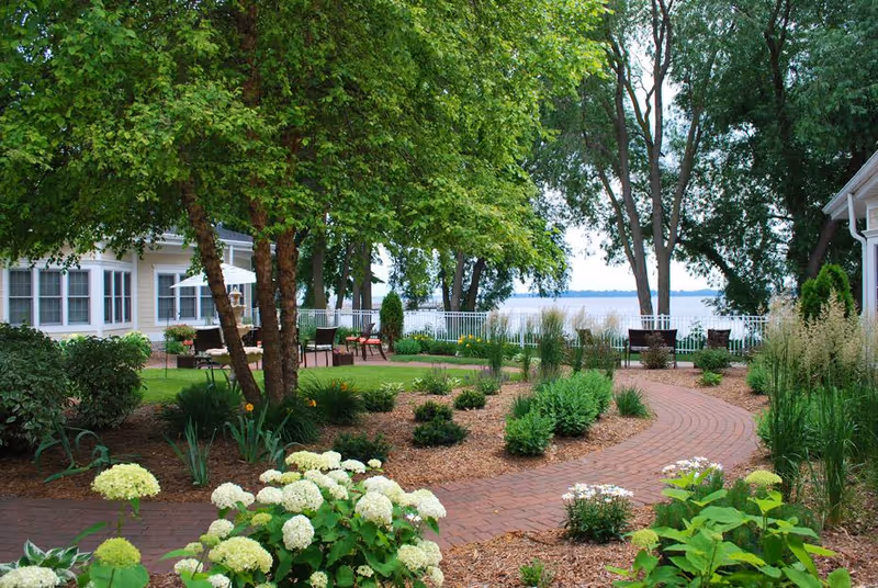 A landscaped outdoor garden area with a brick pathway winding through green shrubs, flowering plants, and trees. There are benches and chairs near a white fence overlooking a large body of water in the background. A white building with multiple windows and an umbrella-covered seating area is visible on the left side.