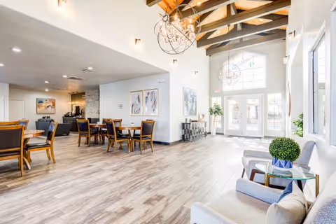 Bright modern senior living common lounge with seating, dining tables, high beamed ceiling, and large entrance windows.