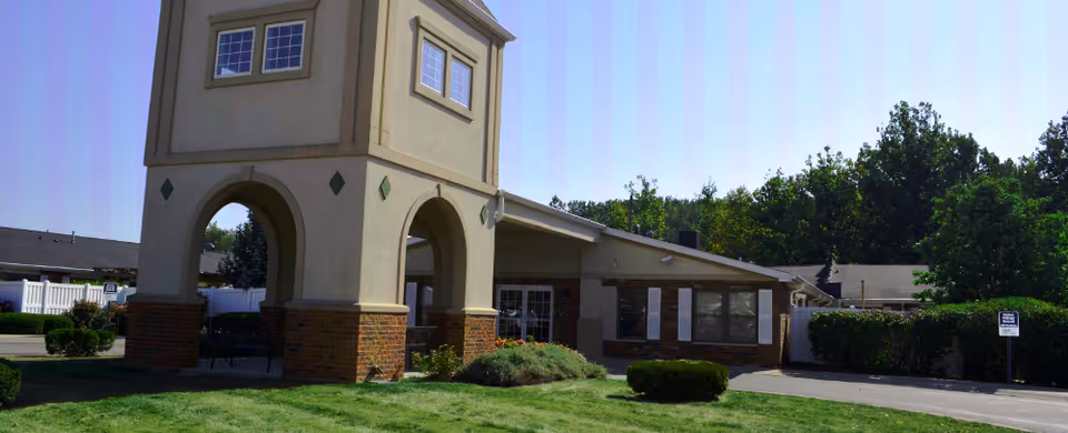 Front exterior of Riverwalk Village showing an arched tower entrance, lawn, driveway and surrounding trees.