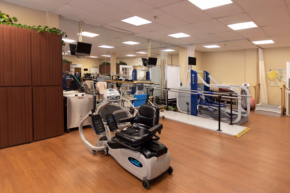 Interior view of a rehabilitation or physical therapy room with exercise equipment including a recumbent bike and an anti-gravity treadmill. The room has wood flooring, beige walls, ceiling lights, and some plants on top of cabinets. There are also privacy curtains and several monitors mounted on poles.