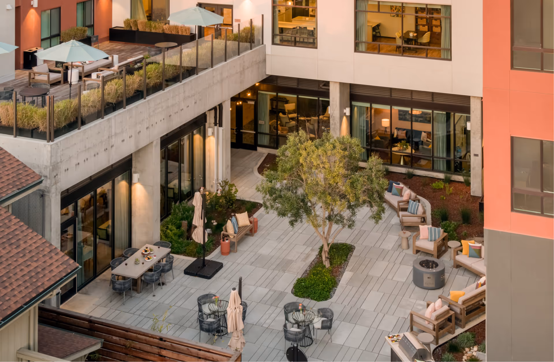 Outdoor courtyard area of a senior living facility with seating arrangements including sofas, armchairs with colorful cushions, round tables with chairs, umbrellas, and a central tree surrounded by greenery. The courtyard is adjacent to a building with large windows showing interior rooms with furniture.