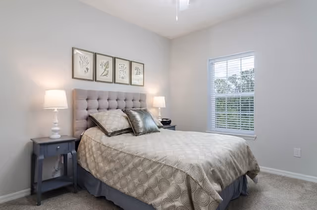 A neatly made bed with a tufted headboard in a bedroom featuring two bedside tables with lamps, a window with blinds, and four framed botanical prints on the wall above the bed.