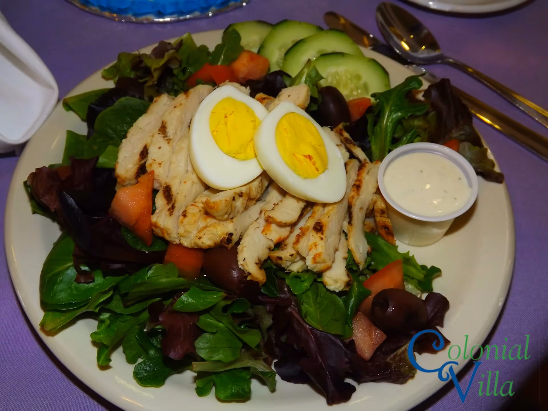 A plate of salad with grilled chicken strips, two halves of a hard-boiled egg, cucumber slices, olives, and mixed greens, served with a small cup of creamy dressing on the side. The plate is on a purple tablecloth with a fork and spoon nearby.