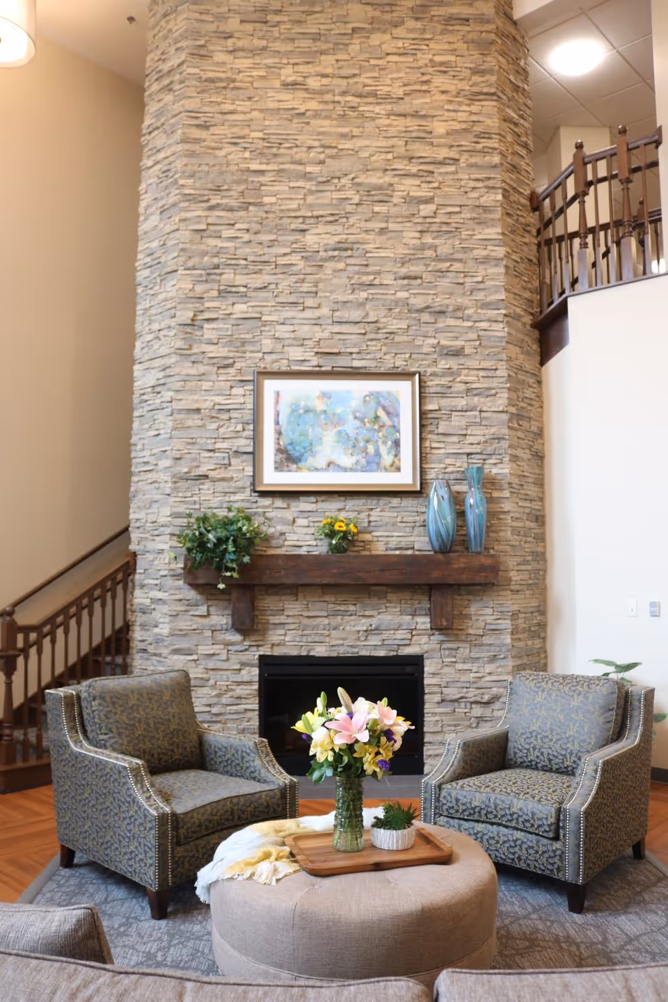 A cozy sitting area in a senior living facility featuring two patterned armchairs facing a round ottoman with a wooden tray holding a vase of colorful flowers and a small potted plant. Behind the chairs is a tall stone fireplace with a wooden mantel decorated with two blue vases, a small plant, and a framed abstract painting. A staircase with wooden railing is visible to the left and right of the fireplace.