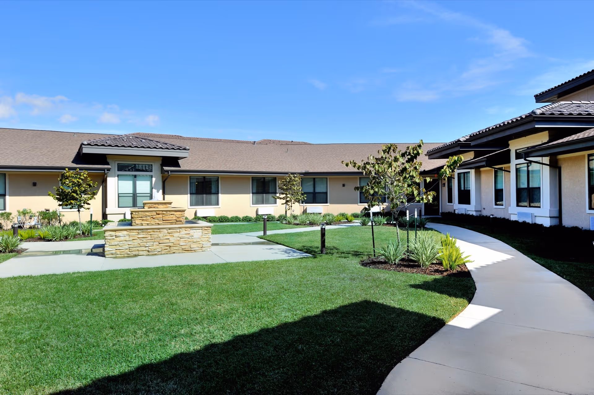 Courtyard of a senior living facility with a stone water feature, green lawn, walkway and single-story buildings under a blue sky.