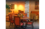 Round dining table with a floral centerpiece surrounded by chairs in a warm-lit common area with armchairs and decorative wall art.