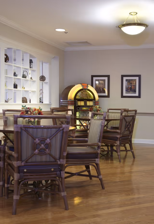 A cozy dining area with wooden tables and cushioned chairs arranged on a polished hardwood floor. A vintage jukebox is positioned against the wall, flanked by two framed pictures. A decorative shelving unit with various ornaments is visible through an open wall partition.