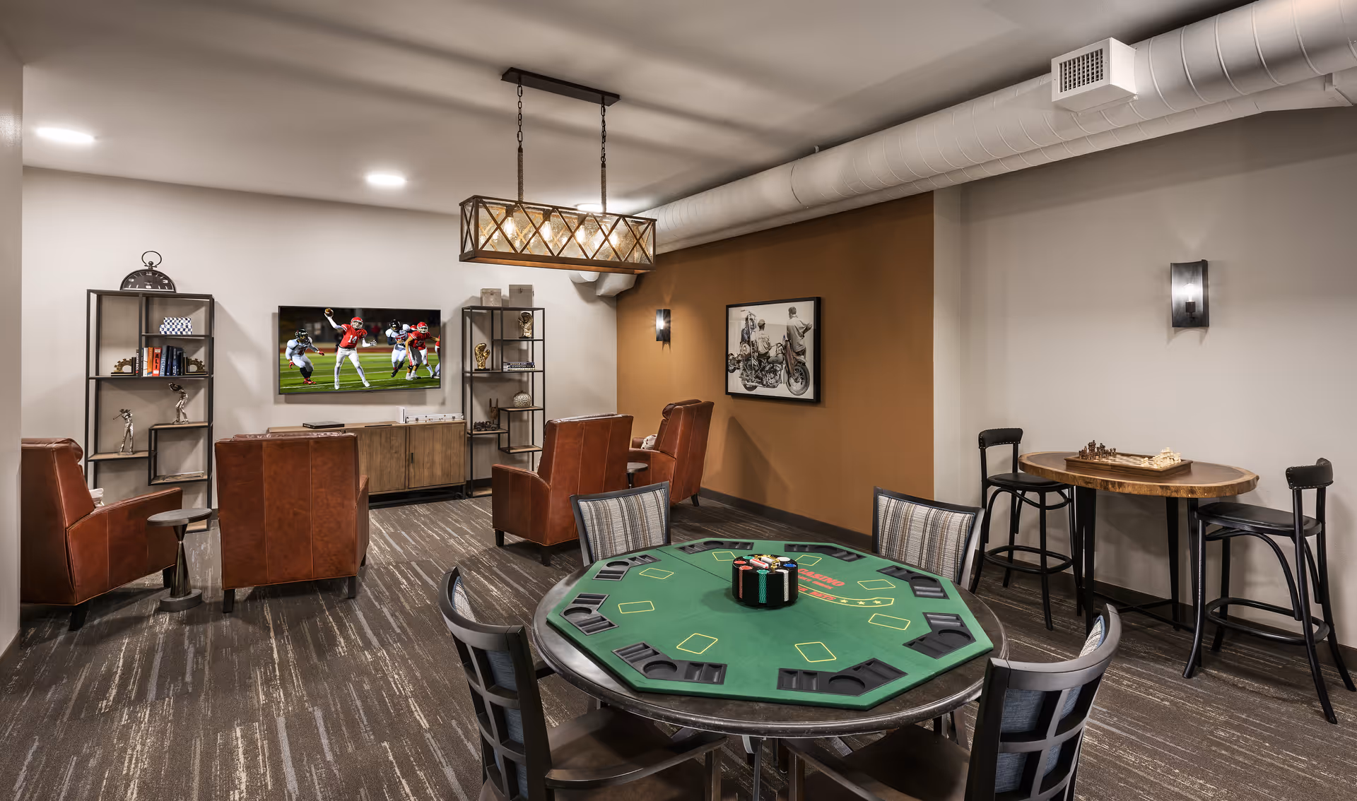 A cozy common area with a poker table in the foreground, surrounded by chairs. In the background, there are four brown leather armchairs facing a wall-mounted TV showing a football game. Two metal shelving units flank the TV, holding decorative items and books. To the right, a small round table with two chairs has a chess set on it. The room has modern lighting and a warm, inviting atmosphere.