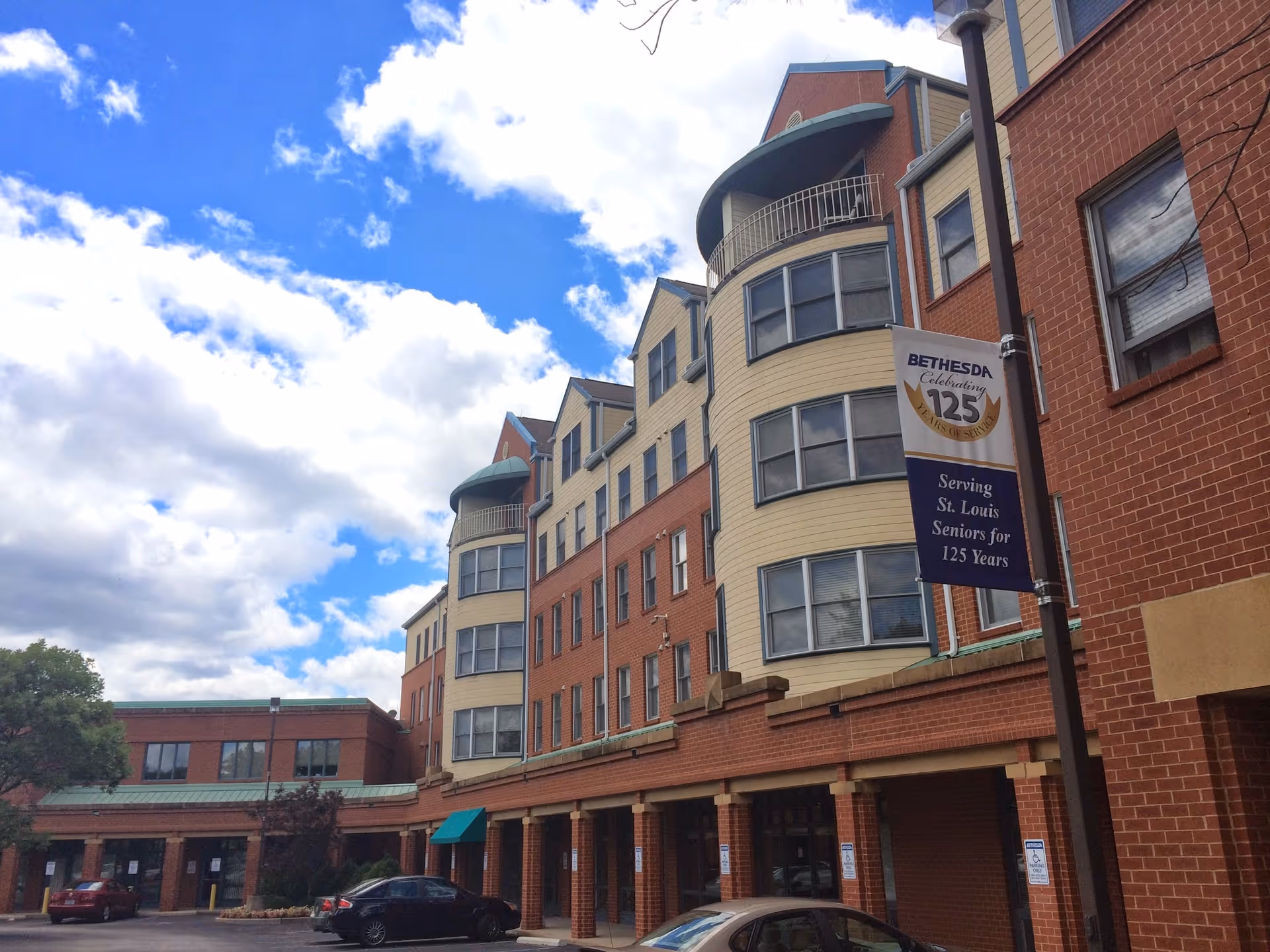 Front exterior of a multi-story red brick senior living building with rounded bay windows, a banner reading 'Bethesda', parked cars, and a partly cloudy sky.