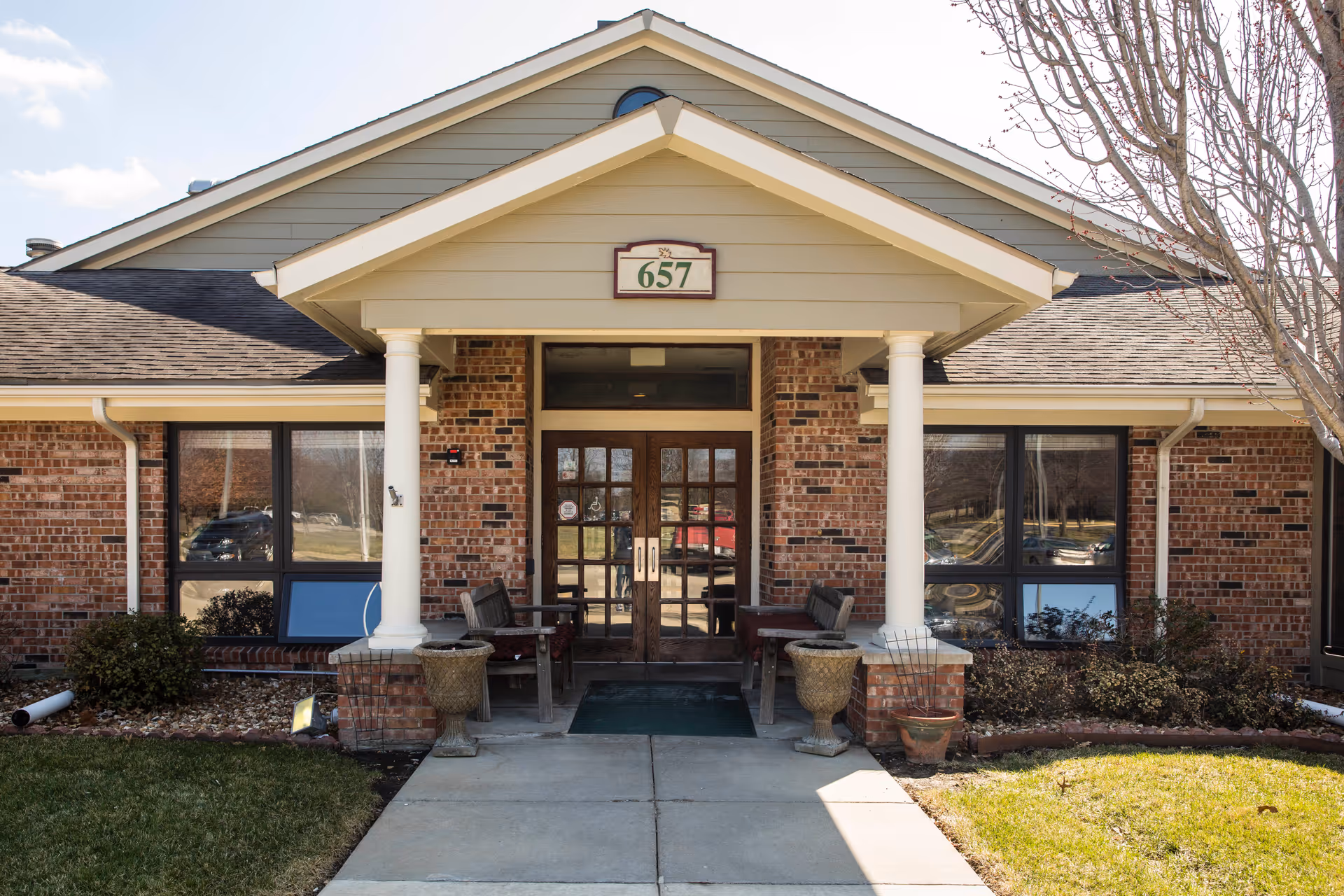 Front entrance of a brick building with a peaked roof and two white columns supporting a small porch. There are two wooden benches and large planters on either side of the entrance door. The building number 657 is displayed above the door. Leafless trees and trimmed bushes are visible around the entrance.