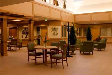 Indoor senior living lounge with round tables, chairs, and potted plants in a spacious lobby.