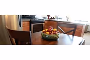 Dining area with a wooden table, chairs, and a fruit bowl near kitchen cabinets.