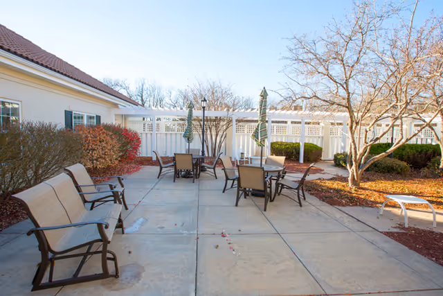 Sunlit outdoor patio with several tables, chairs, umbrellas and benches surrounded by landscaping and a white fence.