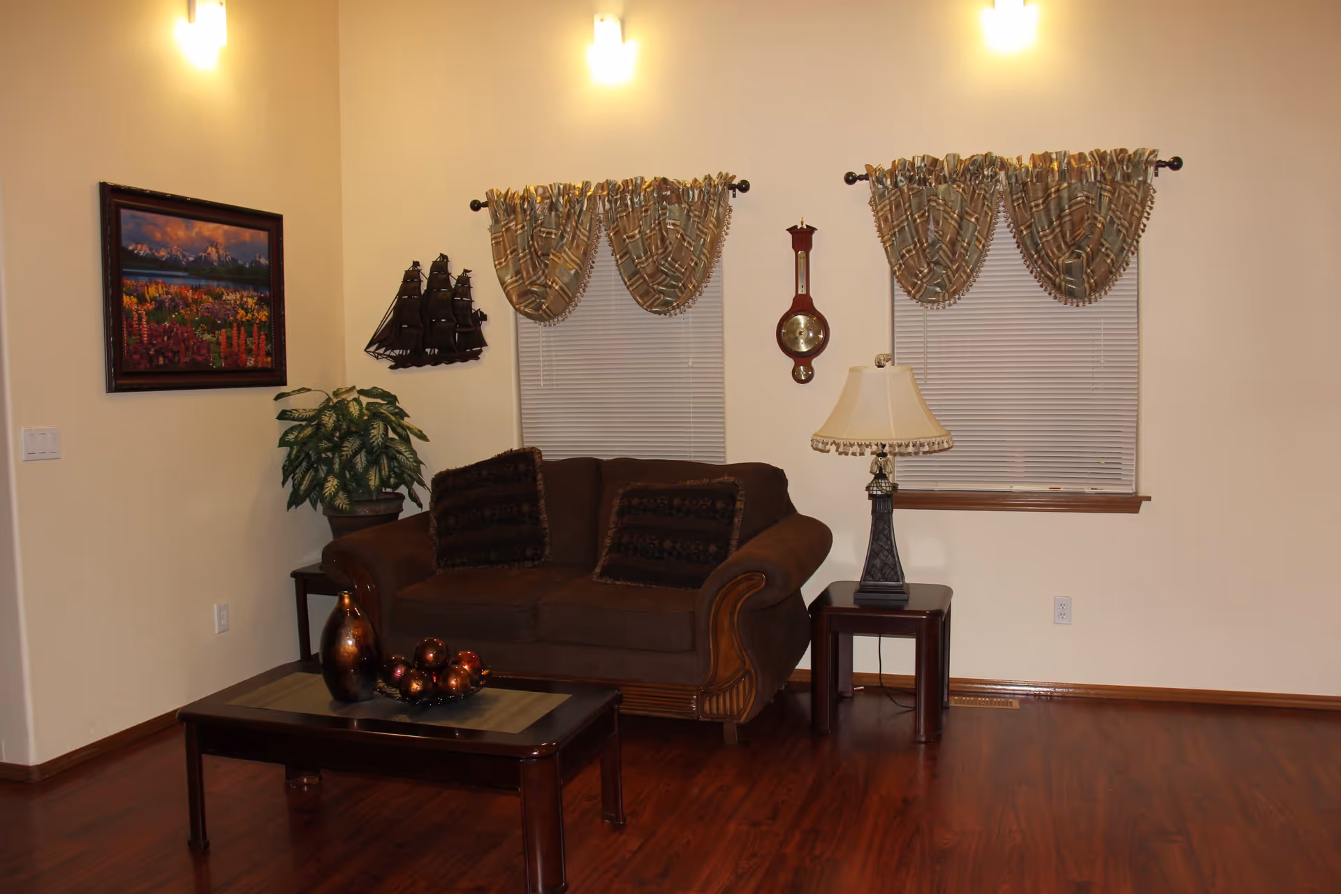 A cozy living room with a brown couch adorned with two dark patterned pillows, a wooden coffee table with decorative items, a side table with a lamp, two windows with patterned curtains, a wall clock, a framed landscape painting, and a potted plant in the corner. The room has wooden flooring and soft wall lighting.