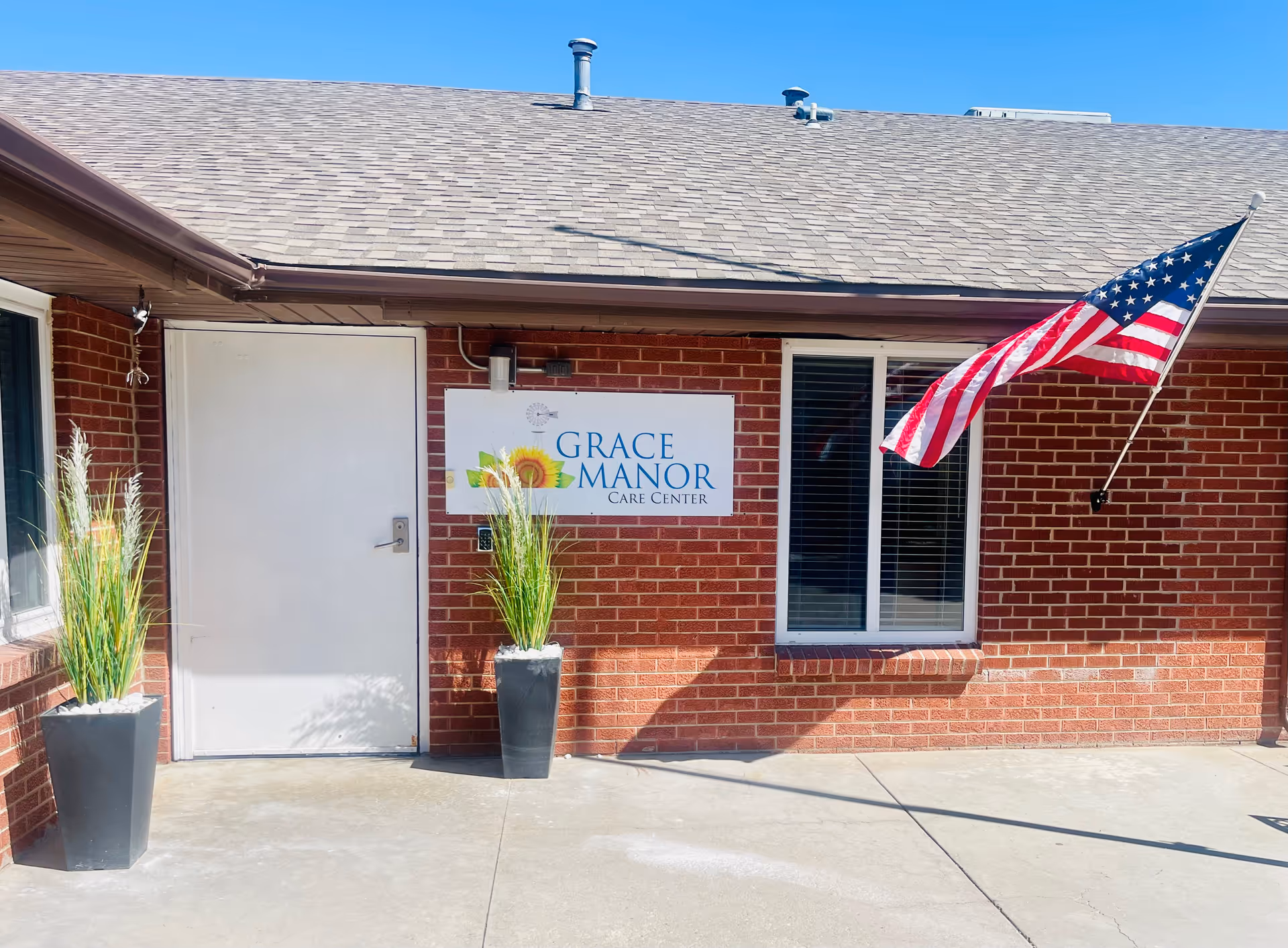 Exterior view of Grace Manor Care Center showing a brick wall with a white door, two tall planters with green plants, a window with blinds, and an American flag mounted on the wall.