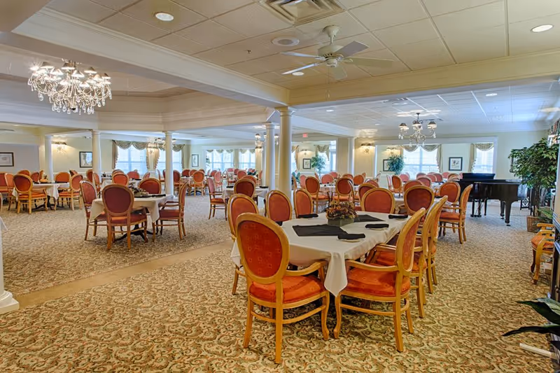 A spacious dining room with multiple round and rectangular tables covered with white tablecloths and set with black napkins. The chairs have wooden frames with red cushioned seats and backs. The room features patterned carpet, chandeliers, ceiling fans, large windows with curtains, and a grand piano in the background. There are decorative plants and framed artwork on the walls.