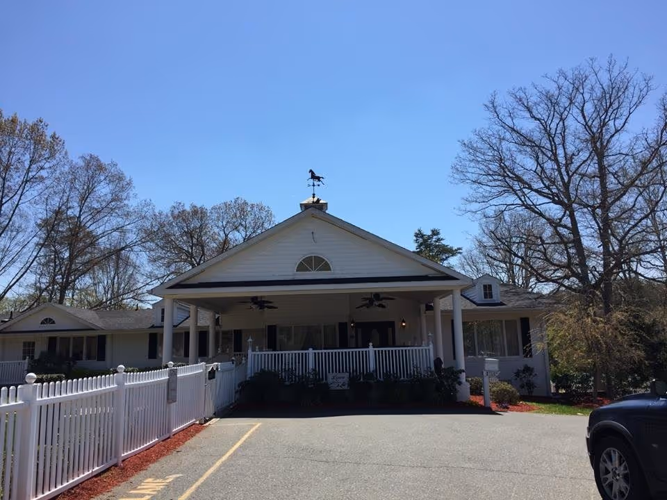 Front exterior view of a single-story building with a white picket fence, a covered porch with ceiling fans, and a weather vane on the roof. Trees surround the building under a clear blue sky.