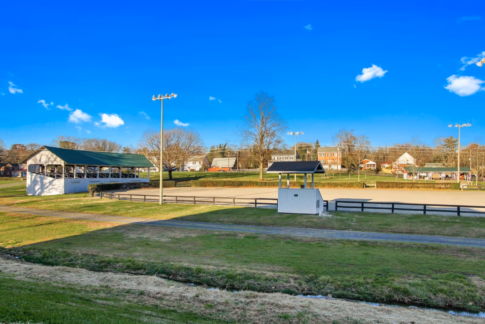 Outdoor view of a large open field with a fenced sandy area, light poles, a small covered structure, and a larger covered pavilion. Trees and houses are visible in the background under a clear blue sky.