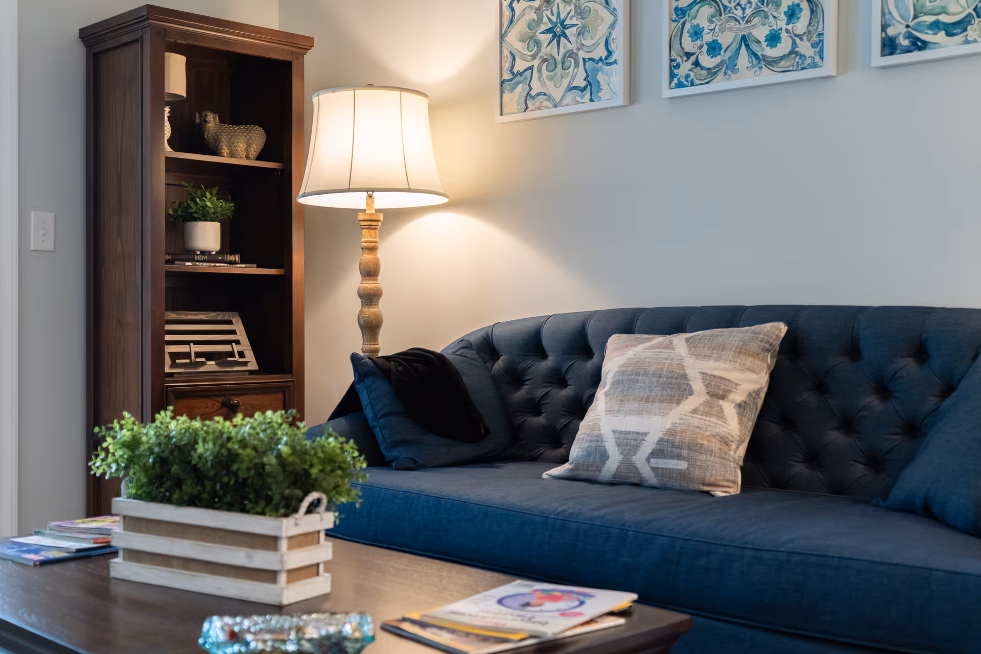 A cozy living room area with a dark blue tufted sofa adorned with patterned and solid pillows. Next to the sofa is a wooden floor lamp with a white lampshade, casting warm light on the wall. Behind the lamp is a wooden bookshelf with decorative items including a small plant and a sculpture. On the coffee table in front of the sofa, there is a rectangular wooden planter with green plants and some magazines or books.