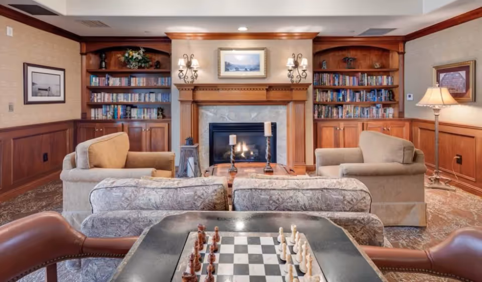 A cozy living room with a fireplace in the center, flanked by built-in wooden bookshelves filled with books. Two beige armchairs face the fireplace, with a patterned sofa behind them. In the foreground, there is a table with a chessboard and chess pieces set up. The room has warm wood paneling and soft lighting from wall sconces and a floor lamp.