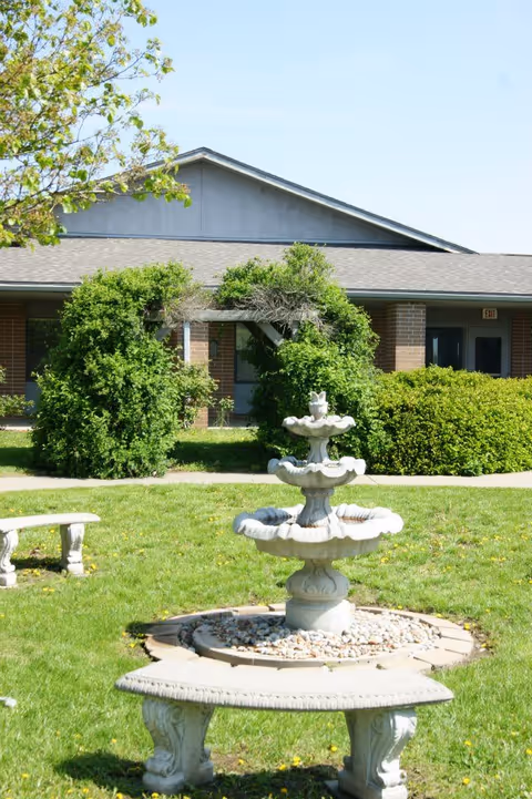 Outdoor garden area with a three-tiered stone fountain surrounded by a circular stone border filled with small rocks. In front of the fountain is a curved stone bench. Behind the fountain, there is a green archway covered with leafy bushes and a building with brick columns and a gray roof in the background under a clear blue sky.