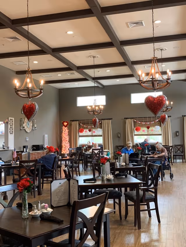 A dining room in a senior living facility decorated with red heart-shaped balloons and Valentine's Day decorations. Several elderly residents are seated at dark wooden tables with chairs, some engaged in conversation or activities. The room has large windows with beige curtains, wooden flooring, and chandeliers hanging from a coffered ceiling. Tables are set with menus, napkins, and small vases with red flowers.