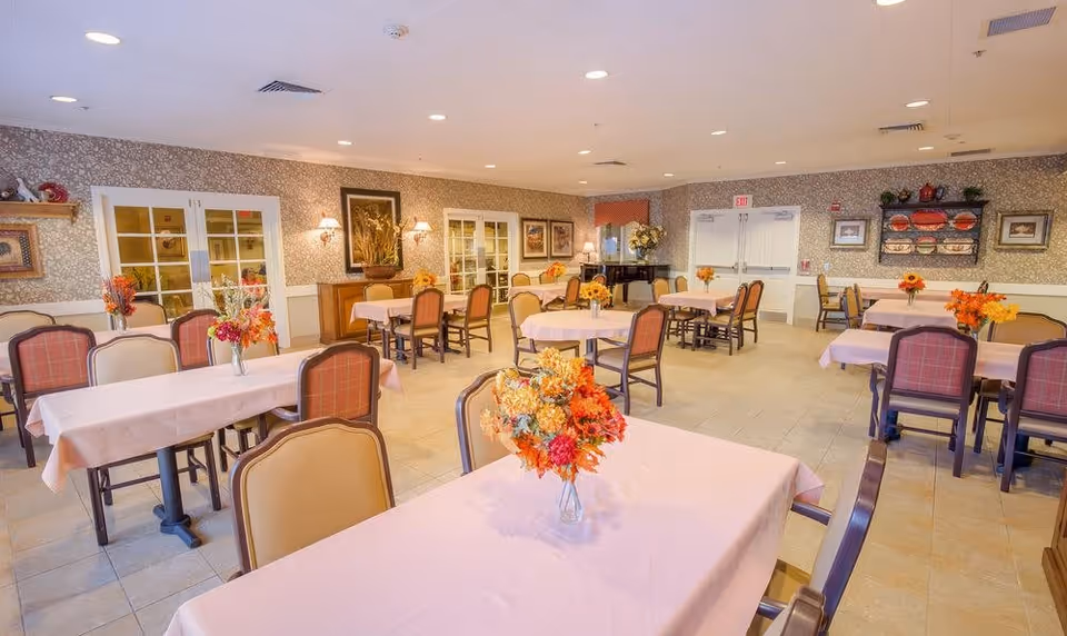 A spacious dining room with multiple tables covered in light pink tablecloths, each adorned with a vase of colorful flowers. The room has beige tiled floors, patterned wallpaper, framed artwork on the walls, and soft lighting from wall sconces and ceiling lights. There are several chairs around each table, and a piano is visible in the back corner near double doors.