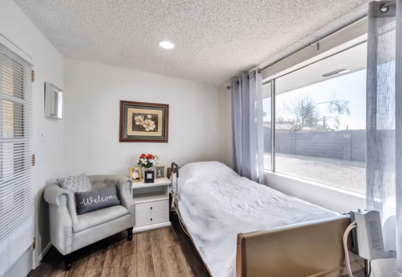 A small, cozy bedroom in an assisted living facility with a single bed covered in a white blanket next to a large window with sheer curtains. There is a gray armchair with a 'Welcome' pillow, a white nightstand with framed photos and a vase of flowers, and a framed floral painting on the wall.