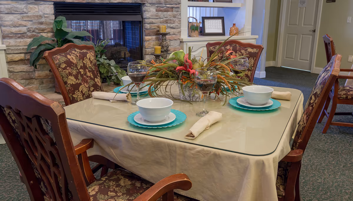 A dining table set for four with floral upholstered wooden chairs, white bowls on turquoise plates, beige napkins, and wine glasses with red wine. The table has a beige tablecloth and a glass top. In the background, there is a stone fireplace with plants and candles, and a doorway leading to another room.