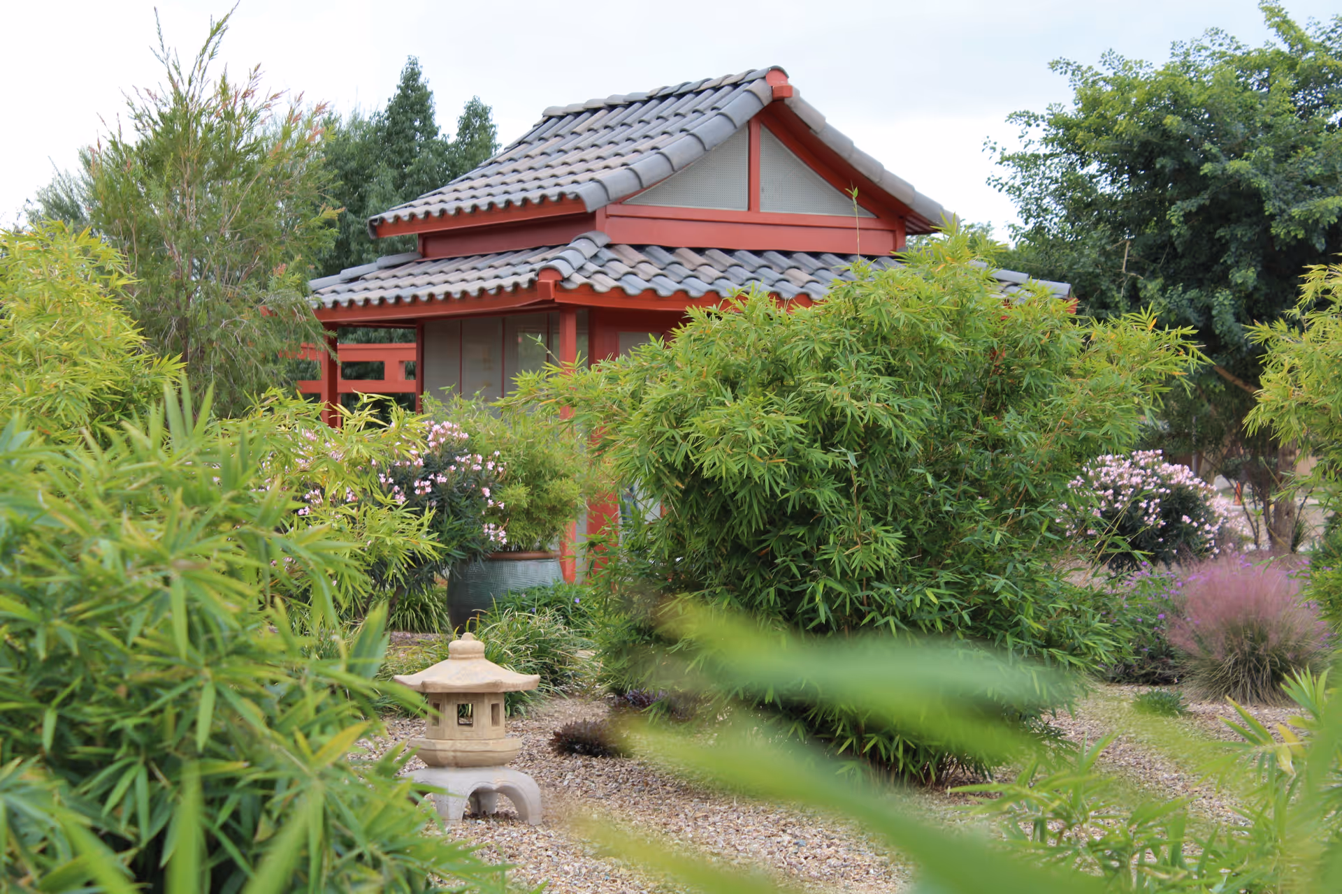 A serene garden area with lush green bushes and plants surrounding a small structure with a tiled roof and red wooden beams, resembling a Japanese-style gazebo. There is also a stone lantern in the foreground, and trees in the background under a cloudy sky.