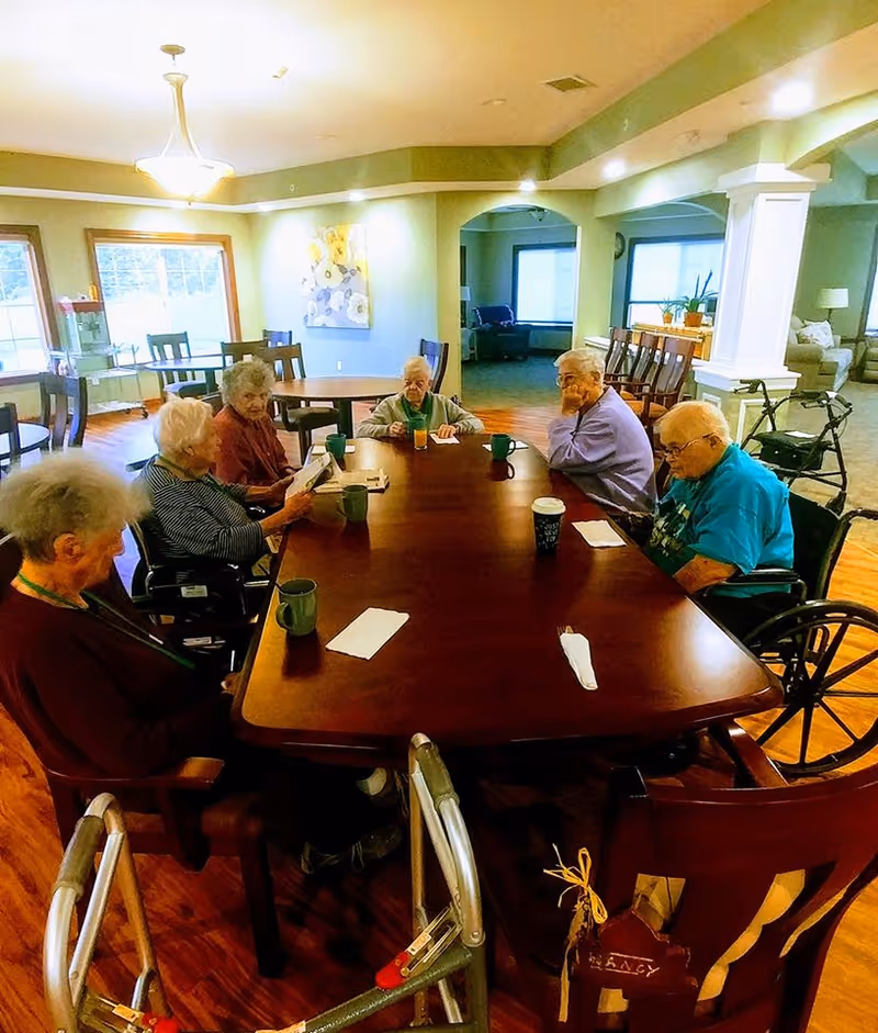 A group of elderly individuals sitting around a large wooden table in a well-lit common area of an assisted living facility. Some are in wheelchairs, and there are cups and napkins on the table. The room has wooden flooring, large windows, and comfortable seating in the background.