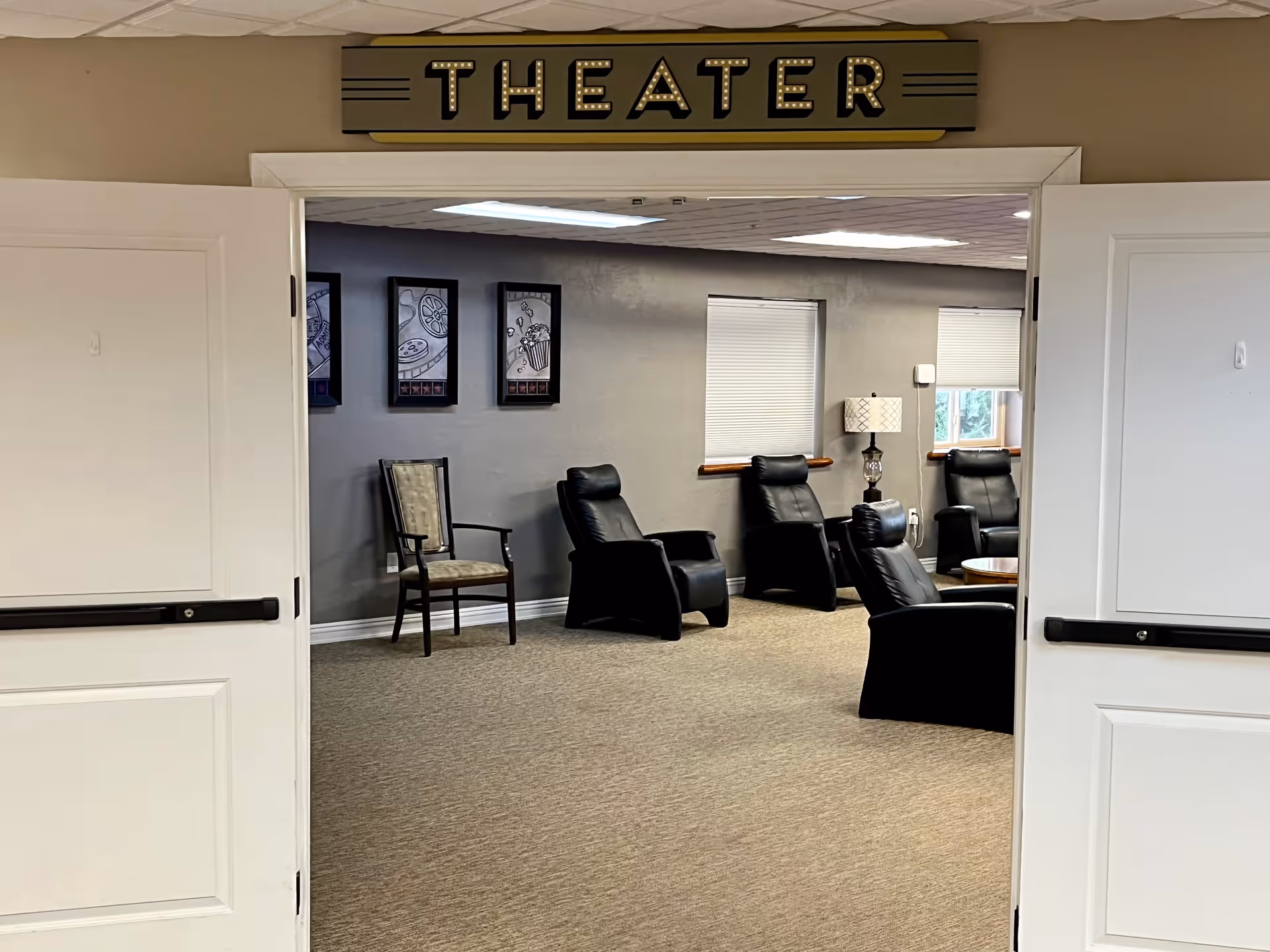 Double doors under a 'THEATER' sign open into a seating room with black recliner chairs, a lamp, framed art, and windows.