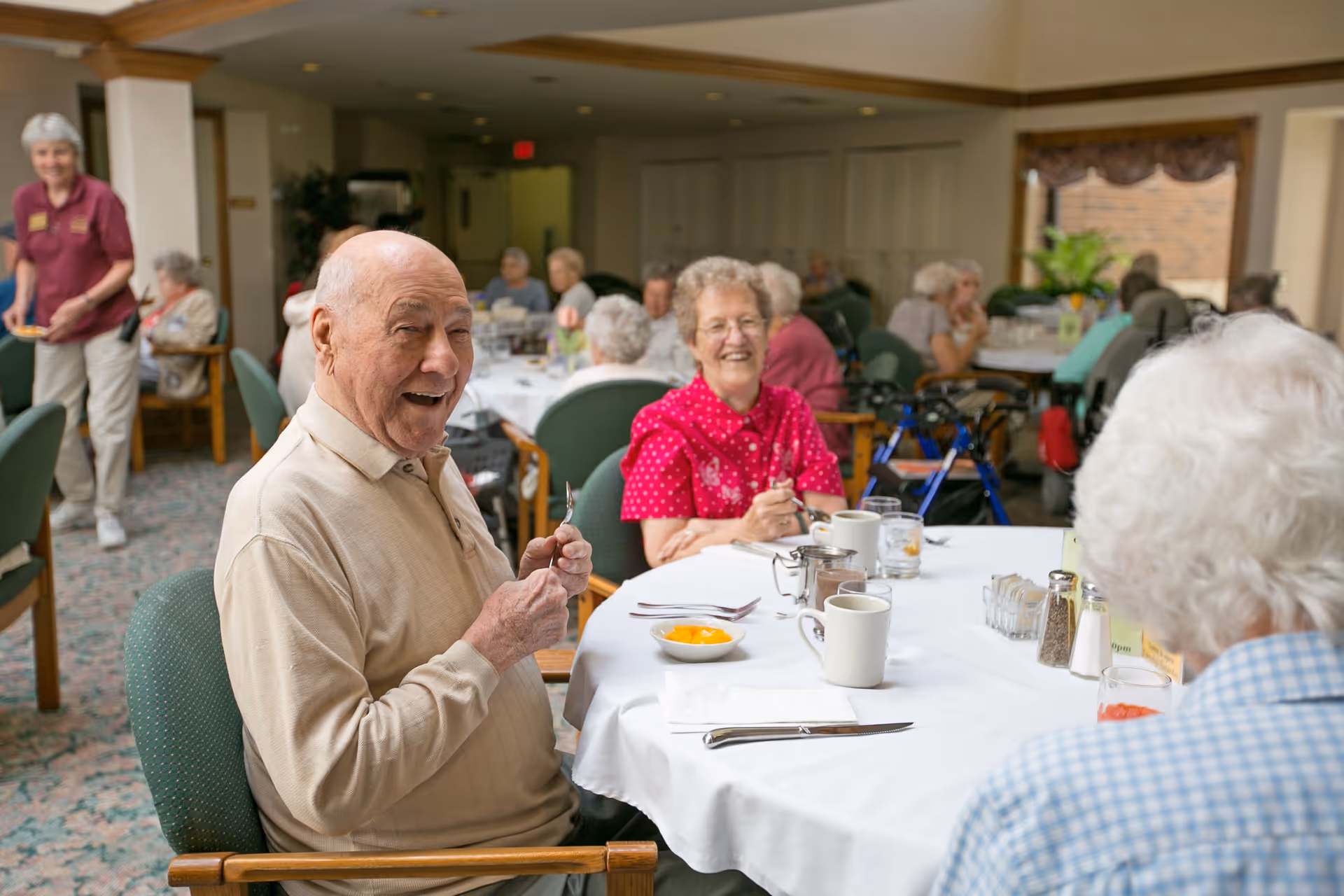 A group of elderly people sitting around a table in a dining room at a senior living facility, smiling and enjoying a meal together. The table is set with cups, utensils, and a bowl of fruit. Other residents and a staff member are visible in the background.
