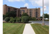 Exterior view of a multi-story brick building with several windows, surrounded by well-maintained green lawns and bushes. A concrete walkway leads towards the building entrance under a partly cloudy sky. A flagpole with a flag is visible on the right side of the image.
