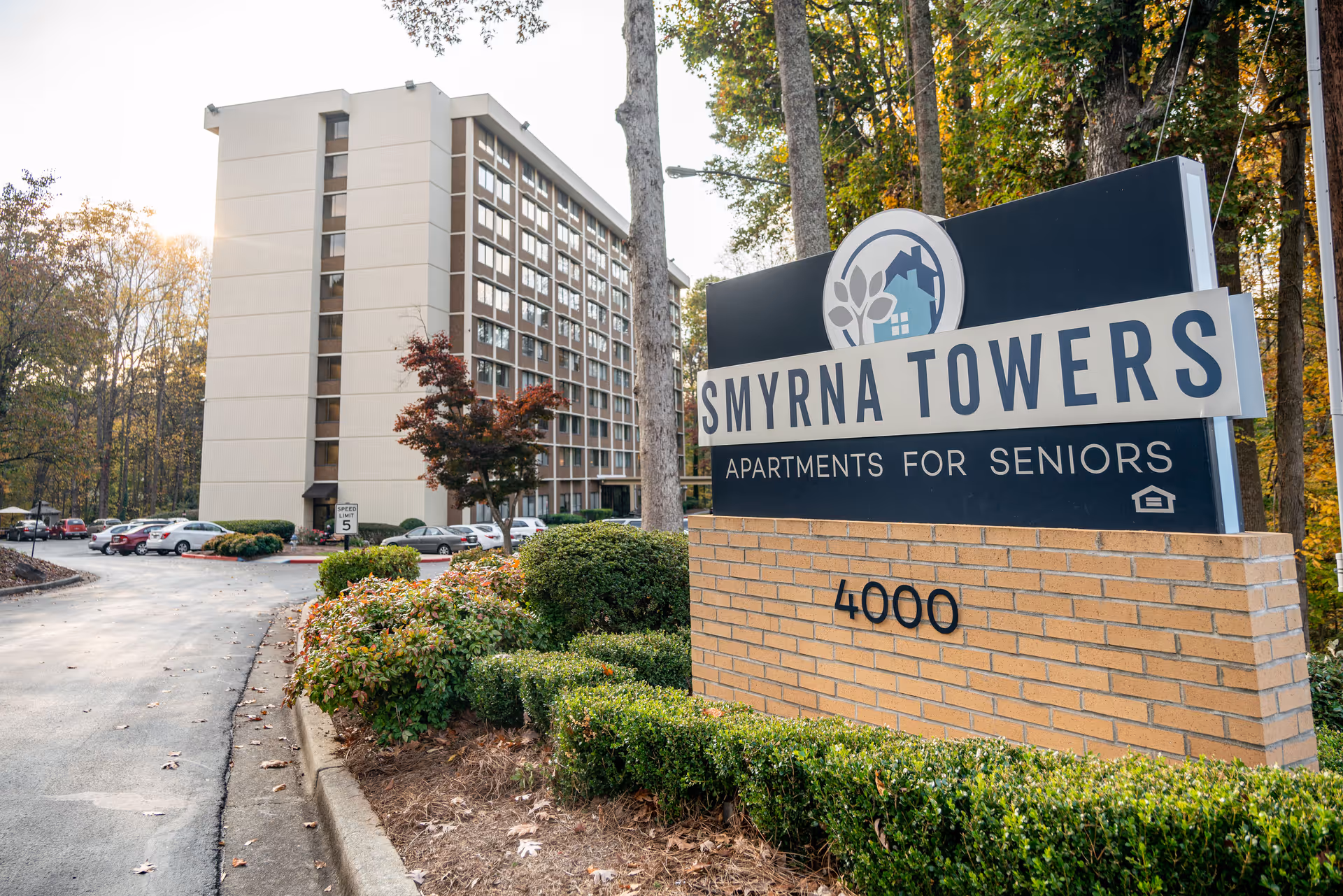 Exterior view of Smyrna Towers Senior Apartments building with a large sign in the foreground that reads 'Smyrna Towers Apartments for Seniors' and the number 4000. The building is surrounded by trees and a parking lot with several cars.