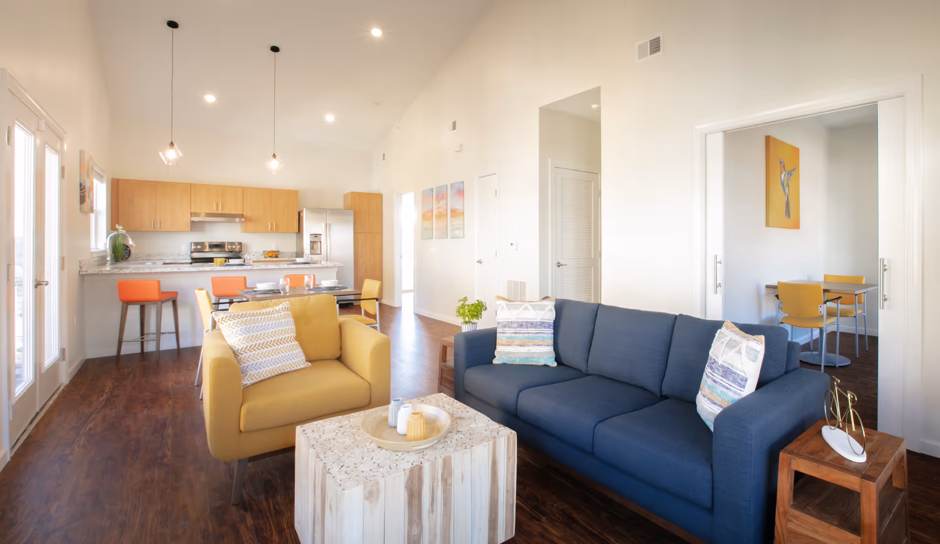 Bright and modern open-concept living area featuring a blue sofa with patterned pillows, a yellow armchair, a wooden coffee table with decorative items, and a small wooden side table with a bicycle sculpture. In the background, there is a kitchen with light wood cabinets, a stainless steel refrigerator, stove, and a breakfast bar with orange stools. Adjacent to the living area is a dining table with yellow chairs and a colorful bird painting on the wall.