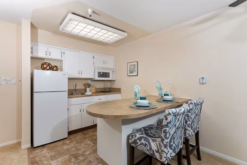 A small kitchen area with white cabinets, a white refrigerator, a microwave, and a sink. There is a countertop island with two patterned upholstered chairs and place settings for two, including plates, bowls, cups, and folded napkins. The walls are beige and there is a framed picture hanging on the wall.