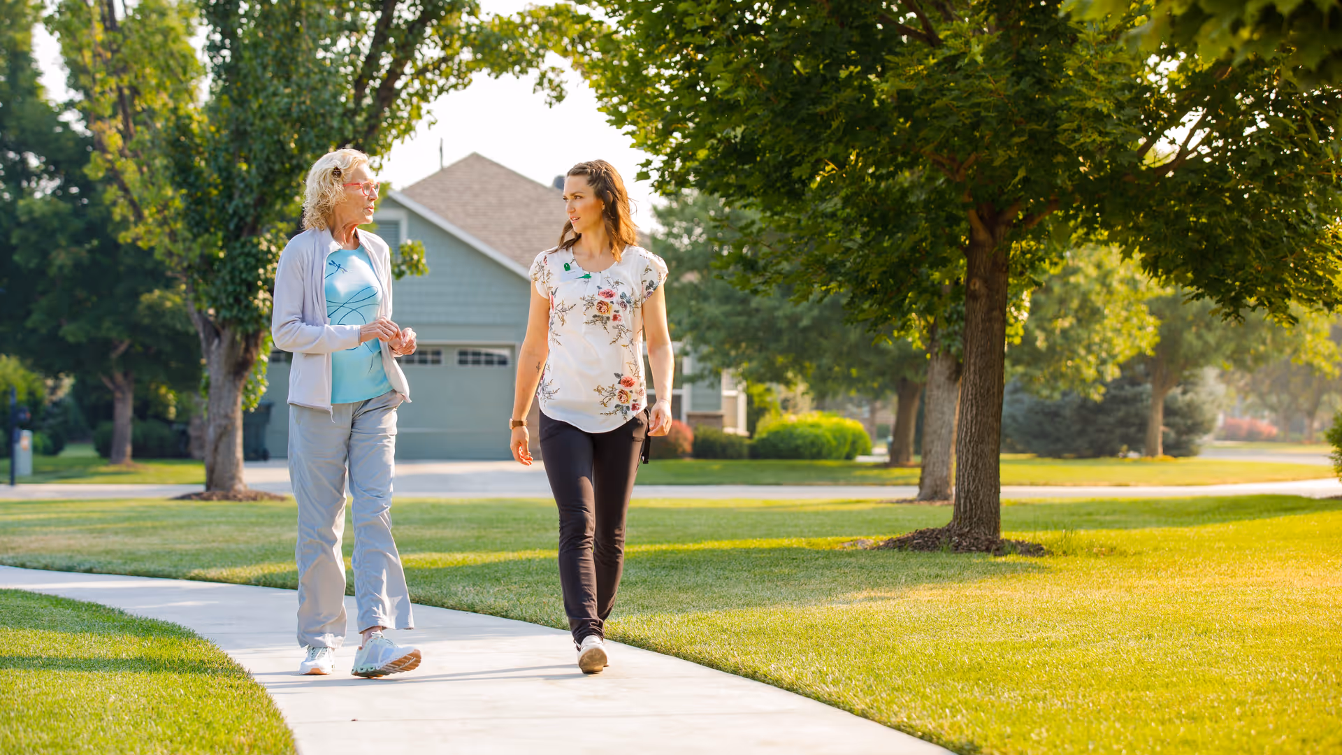 An elderly woman and a younger woman walking and talking on a sidewalk in a residential neighborhood with green lawns and trees on a sunny day.