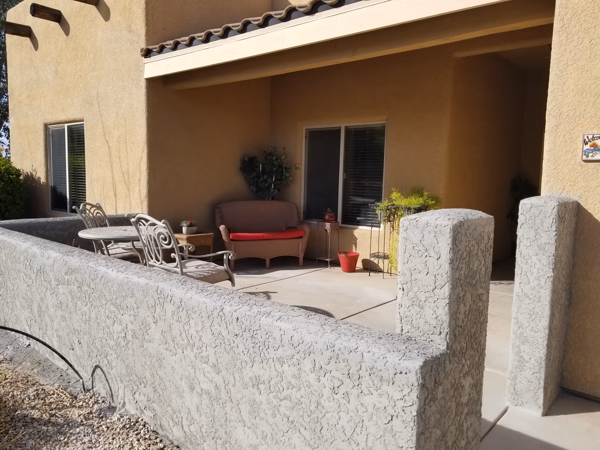 Outdoor patio area with a beige stucco wall and floor, featuring a small round table with two metal chairs, a wicker loveseat with a red cushion, several potted plants, and a window with closed blinds. The patio is partially shaded by the building's roof.