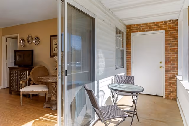 Covered patio with a small round glass table and two wicker chairs outside a sliding glass door into a living area.