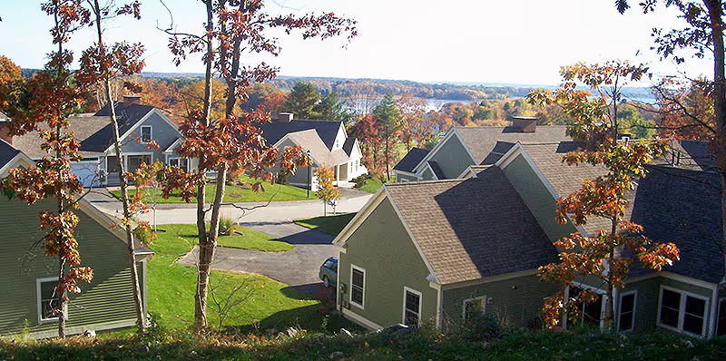 View of a residential community with multiple houses surrounded by trees with autumn foliage, green lawns, and paved roads under a clear sky.