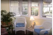 A cozy sunroom with large windows letting in natural light, featuring two white wicker chairs with blue cushions, a small side table with a lamp and decorative figurine, and a potted plant on a stand.