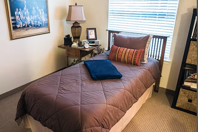 A neatly made single bed with a brown quilt, a blue folded blanket, and two pillows, positioned next to a window with blinds. Beside the bed is a wooden nightstand with a lamp, framed photo, and decorative items. A framed picture hangs on the wall above the nightstand, and a shelving unit with baskets is visible on the right side of the image.