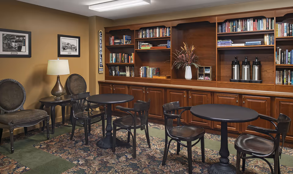 Cozy common room with round tables and chairs, wooden bookshelves, and a beverage station.