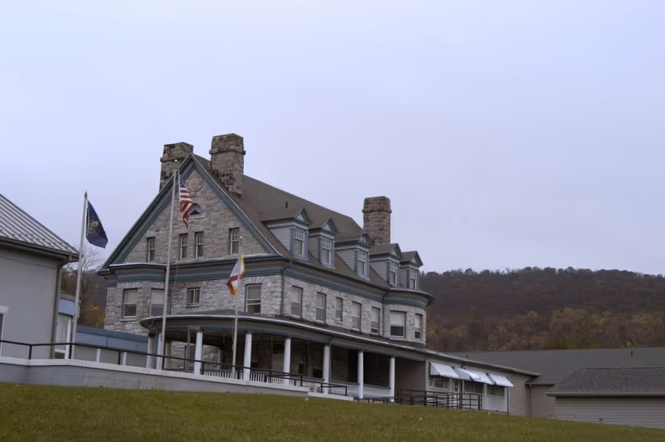 A large stone building with multiple chimneys and dormer windows, featuring a covered porch with white columns. Three flagpoles with flags are in front of the building, and a grassy hill slopes down in the foreground. Trees and hills are visible in the background under a cloudy sky.